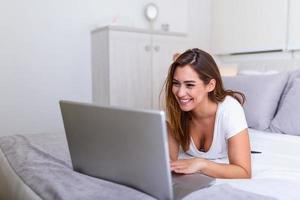 schöne junge College-Studentin, die zu Hause Hausaufgaben im Bett macht, junge hübsche Frau bereitet Schulprüfung im Schlafzimmer mit Laptop-PC vor. freiberufliches arbeiten von zu hause aus foto