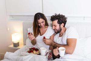 Süßes Paar beim Frühstück im Bett im Schlafzimmer. Schöne Frau, die ihrem Freund Erdbeeren im Bett füttert, während sie im Schlafzimmer frühstückt und Kaffee trinkt foto