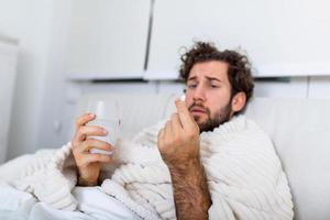 Konzept des Leidens unter schrecklichen Schmerzen. Schöner, trauriger, verärgerter, unglücklicher, beunruhigter, schwacher Mann mit lockigem Haar, unter der Decke liegt im Bett und nimmt eine Pille foto