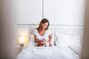 Attraktive Frau im Schlafanzug, die morgens mit dem Geruch von Kaffee und frischem Frühstück auf einem Holztablett im Bett aufwacht foto