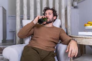 Nach der Arbeit sitzt ein depressiver Typ auf dem Sofa und trinkt ein kaltes Bier. Hand hält eine Flasche, Mann trinkt allein nach Hause. alkoholismus, alkoholsucht und menschenkonzept foto