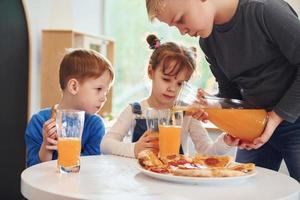 Drei Kinder sitzen drinnen am Tisch und essen gemeinsam Pizza mit Orangensaft foto