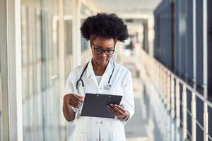 Junge afroamerikanische Ärztin in weißer Uniform mit Stethoskop und Notizblock im Korridor foto