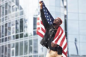 patriot, der usa-flagge hält. Vorstellung von Stolz und Freiheit. junger afrikanisch-amerikanischer Mann in schwarzer Jacke im Freien in der Stadt, der gegen ein modernes Geschäftsgebäude steht