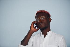 Junger afroamerikanischer Mann in weißer formeller Kleidung, der drinnen an der Wand telefoniert foto