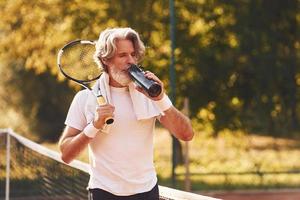 Pause machen und Wasser trinken. Älterer stilvoller Mann in weißem Hemd und schwarzen sportlichen Shorts auf dem Tennisplatz foto