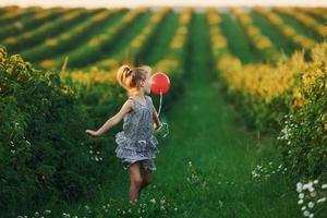 positives kleines mädchen mit rotem ballon in den händen hat spaß auf dem feld zur sommertageszeit foto
