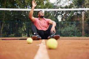 sitzt in der Nähe des Netzes und macht eine Pause. afroamerikanischer mann im rosa hemd sitzt mit tennisschläger auf dem platz im freien foto