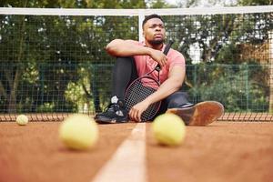 sitzt in der Nähe des Netzes und macht eine Pause. afroamerikanischer mann im rosa hemd sitzt mit tennisschläger auf dem platz im freien foto