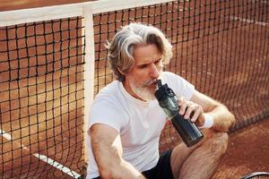 Wasser trinken und auf dem Boden sitzen. Älterer, moderner, stilvoller Mann mit Schläger im Freien auf dem Tennisplatz tagsüber foto