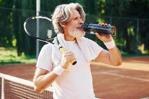 älterer moderner stilvoller mann mit schläger draußen auf tennisplatz tagsüber trinkwasser foto