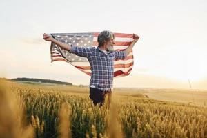 usa-flagge in den händen halten. patriotischer älterer stilvoller mann mit grauem haar und bart auf dem landwirtschaftlichen feld foto