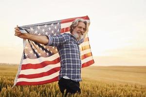 usa-flagge in den händen halten. patriotischer älterer stilvoller mann mit grauem haar und bart auf dem landwirtschaftlichen feld foto