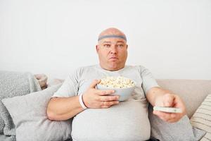 sitzt mit Popcorn auf dem Sofa. lustiger übergewichtiger mann in lässiger kleidung ist drinnen zu hause foto