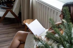 frau im warmweißen winterpullover sitzt neben dem fenster zu hause am heiligabend und liest ein buch foto