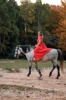 reiten auf einem pferd, wandern in einem herbstwald, eine frau, die auf einem pferd in einem langen roten kleid mit bloßen füßen reitet. foto