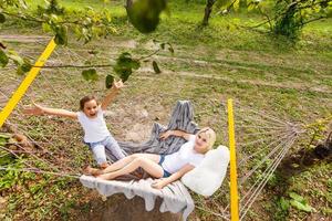 weite Sicht auf eine junge Mutter und Tochter, die sich zusammen entspannen und lächelnd in einer Hängematte sitzen, sich an einem sonnigen Sommertag in einem Ferienhausgarten mit Gras und Bäumen umarmen und entspannen, Lebensstil foto