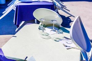 Nahaufnahme eines Restaurants am Strand mit einem weißen Tisch und einem weißen Stuhl vor dem blauen Mittelmeer, serviert mit Tellern und einem Glas und mit einem Seil eingezäunt foto