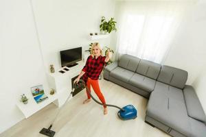 junge lächelnde frau, die den teppich im wohnzimmer staubsaugt, modernes skandinavisches interieur. beschäftigt, Putztag. Zuhause, Haushaltskonzept foto