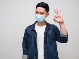 positiver mann jeanshemd tragen medizinische maske hand ok zeichen isoliert foto