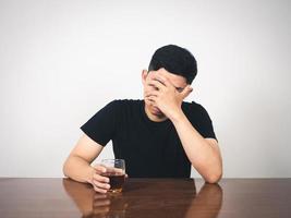 Depressiver Mann, der am Tisch sitzt, schließt sein Gesicht und hält ein Alkoholglas in der Hand foto
