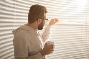 Rückansicht eines jungen, stilvollen Mannes mit Bart und Kaffee, der durch ein Loch in Fensterjalousien späht und auf die Straße blickt. Konzept, die Morgensonne und Positivität zu genießen foto