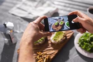 Hände fotografieren auf dem Smartphone zwei schöne, gesunde Sauerrahm- und Avocado-Sandwiches, die an Bord auf dem Tisch liegen. Social Media und Food-Konzept foto