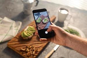 Hände fotografieren auf dem Smartphone zwei schöne, gesunde Sauerrahm- und Avocado-Sandwiches, die an Bord auf dem Tisch liegen. Social Media und Food-Konzept foto