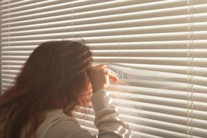 Rückansicht einer schönen jungen Frau mit langen Haaren, die durch ein Loch in den Jalousien blickt und aus dem Fenster schaut. überwachungs- und neugierkonzept foto