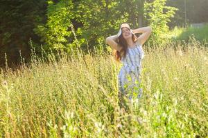 Fröhliche junge Frau mit langen Haaren in Hut und Kleid, die an einem sonnigen Tag durch den Sommerwald geht. sommerfreude konzept foto