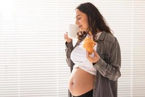 gesunde schöne schwangere frau, die tee trinkt und während des mittagessens croissant isst. konzept der kalorienreichen ernährung während des wartens auf die geburt des babys. Platz kopieren. foto