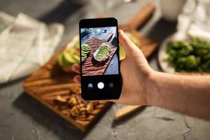 Hände fotografieren auf dem Smartphone zwei schöne, gesunde Sauerrahm- und Avocado-Sandwiches, die an Bord auf dem Tisch liegen. Social Media und Food-Konzept foto