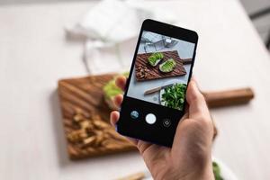 Hände fotografieren auf dem Smartphone zwei schöne, gesunde Sauerrahm- und Avocado-Sandwiches, die an Bord auf dem Tisch liegen. Social Media und Food-Konzept foto