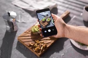 Hände fotografieren auf dem Smartphone zwei schöne, gesunde Sauerrahm- und Avocado-Sandwiches, die an Bord auf dem Tisch liegen. Social Media und Food-Konzept foto