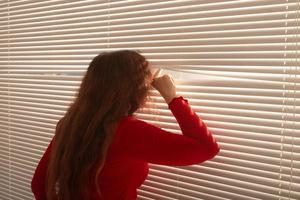 Rückansicht einer schönen jungen Frau mit langen Haaren, die durch ein Loch in den Jalousien blickt und aus dem Fenster schaut. überwachungs- und neugierkonzept foto