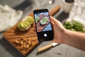 Hände fotografieren auf dem Smartphone zwei schöne, gesunde Sauerrahm- und Avocado-Sandwiches, die an Bord auf dem Tisch liegen. Social Media und Food-Konzept foto