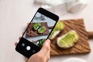 Hände fotografieren auf dem Smartphone zwei schöne, gesunde Sauerrahm- und Avocado-Sandwiches, die an Bord auf dem Tisch liegen. Social Media und Food-Konzept foto