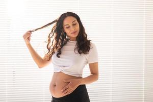 lächelnde junge schöne schwangere Frau, die ihren Bauch und ihr Haar berührt und sich freut. Konzept der Gesundheit und des Nachdenkens über die Zukunft beim Warten auf das Baby. Exemplar foto