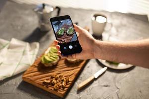 Hände fotografieren auf dem Smartphone zwei schöne, gesunde Sauerrahm- und Avocado-Sandwiches, die an Bord auf dem Tisch liegen. Social Media und Food-Konzept foto
