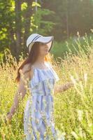 Fröhliche junge Frau mit langen Haaren in Hut und Kleid, die an einem sonnigen Tag durch den Sommerwald geht. sommerfreude konzept foto