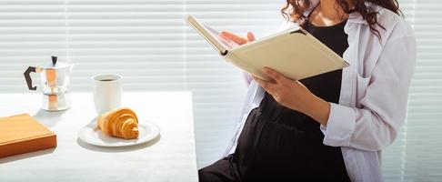 Nahaufnahme einer schwangeren Frau, die zu Hause ein Buch isst und liest. Morgen-, Frühstücks- und Schwangerschaftskonzept foto