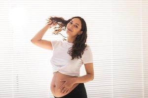 lächelnde junge schöne schwangere Frau, die ihren Bauch und ihr Haar berührt und sich freut. Konzept der Gesundheit und des Nachdenkens über die Zukunft beim Warten auf das Baby. Exemplar foto