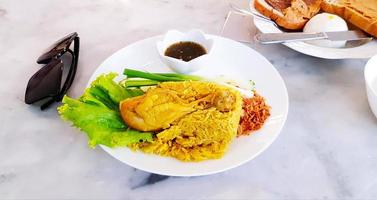 Chicken Biryani mit süßer Soße, getrocknete Zwiebel, frischer Grünkohl, Gurke, Frühlingszwiebeln auf weißem Teller und Suppe auf Schüssel auf Marmortisch mit schwarzer Sonnenbrille und Brot unscharfer Hintergrund. Frühstück foto