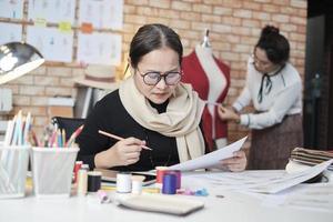 asiatische modedesignerin mittleren alters und junge auszubildende schneiderin arbeiten in einem studio mit bunten fäden und nähstoffen für ideen einer kleiderdesign-kollektion, professionelles boutique-kleinunternehmen. foto