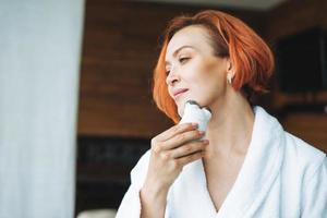 Frau 35 Jahre im weißen Bademantel mit sauberem, frischem Gesicht und Händen mit roten Haaren, die Fasialmassage mit Mikrostrom-Gesichtsmassagegerät im Badezimmer machen, sich selbst verwöhnen, Körperpflege zu Hause foto