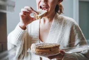 Attraktive, freundliche, brünette Frau mittleren Alters in gemütlicher Strickjacke, die zu Hause Kuchen aus dem Kühlschrank isst foto