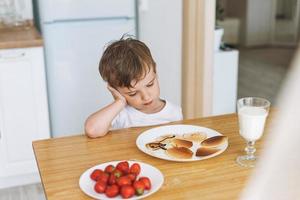 Süßer kleiner Junge, der mit Puncakes mit Beeren in der Küche zu Hause sitzt. Kind will nicht essen foto