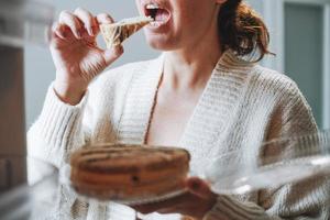 Attraktive, freundliche, brünette Frau mittleren Alters in gemütlicher Strickjacke, die zu Hause Kuchen aus dem Kühlschrank isst foto