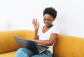 Schöne junge afrikanisch-amerikanische Frau in weißem T-Shirt und Blue Jeans mit Laptop auf gelbem Sofa in hellem, modernem Interieur foto