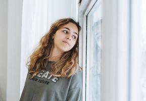 schönes trauriges unglückliches jugendlichermädchen mit dem lockigen haar, das auf der fensterbank sitzt foto